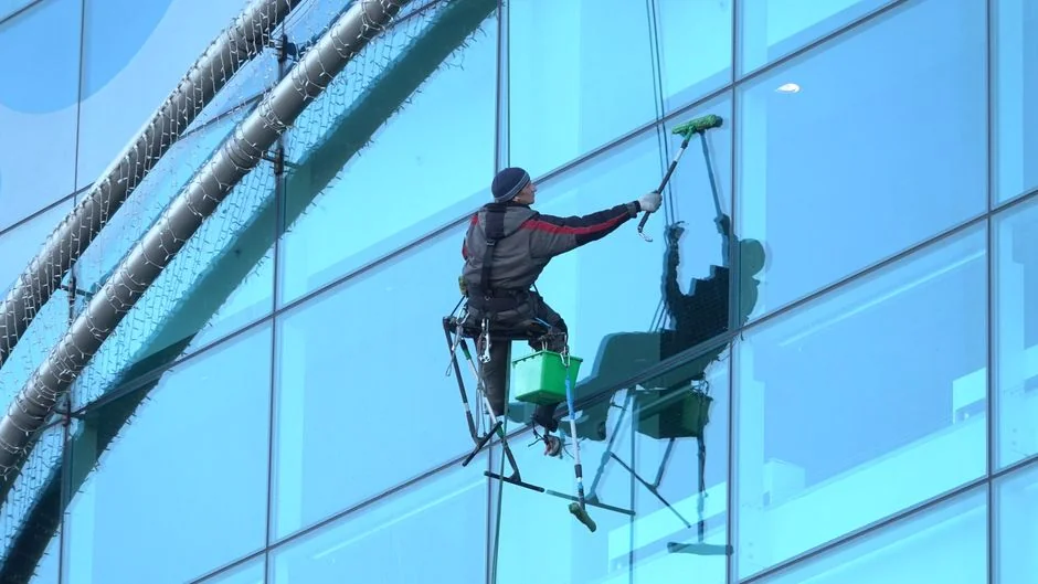 gallery-03 Un homme nettoie les vitres d’un gratte-ciel, harnaché, illustrant le travail en hauteur en milieu urbain.